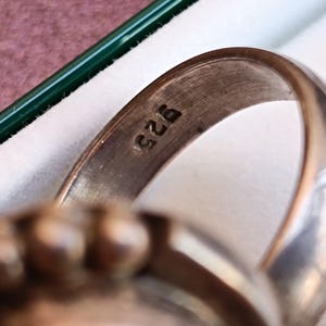 Puede incluir: Primer plano de un anillo de plata con una banda redondeada. El anillo est&aacute; estampado con el n&uacute;mero "925", que indica plata de ley. El dise&ntilde;o del anillo presenta una parte superior texturizada, posiblemente engastada con piedras preciosas. El anillo se muestra en una caja blanca con un borde verde.