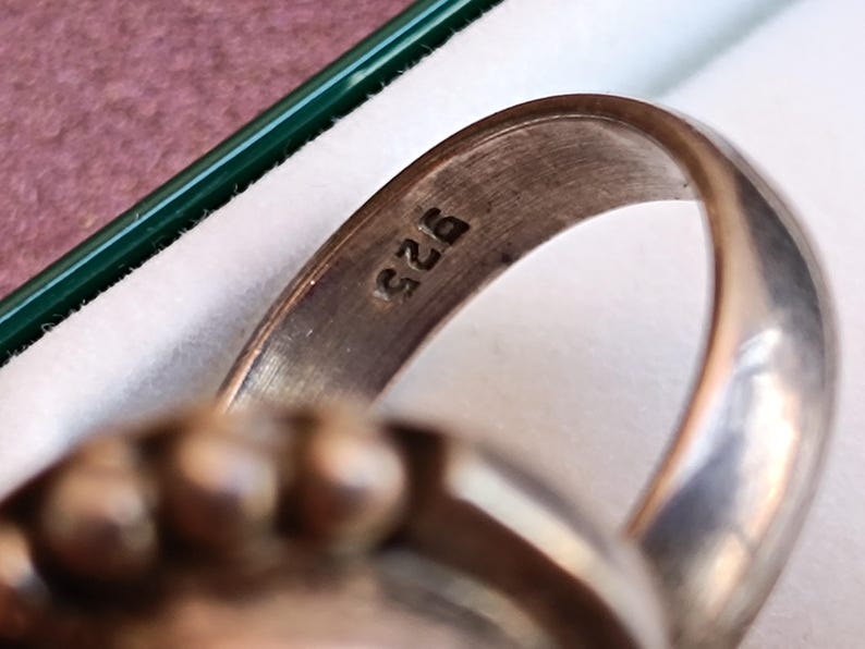 Puede incluir: Primer plano de un anillo de plata con una banda redondeada. El anillo est&aacute; estampado con el n&uacute;mero "925", que indica plata de ley. El dise&ntilde;o del anillo presenta una parte superior texturizada, posiblemente engastada con piedras preciosas. El anillo se muestra en una caja blanca con un borde verde.