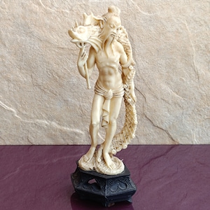 May include: Ivory-colored figurine of a man carrying a dragon. The figure stands on a black, ornate base. The man has a long beard and is draped with a dragon's body. The background is a textured, light brown wall.