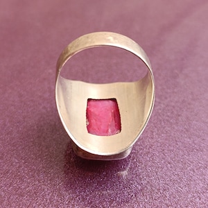May include: A silver ring featuring a rectangular, bright pink gemstone. The ring has a wide, smooth band with a polished finish. The gemstone is the focal point, reflecting light. The ring is set against a purple background.