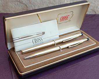 May include: A gold Cross pen and pencil set in a brown and gold gift box. The box has a gold Cross logo with the text "CROSS since 1946".