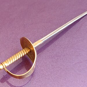 K&ouml;nnte beinhalten: Ein goldfarbener Metall-Cocktail-Stich in Schwertform mit einem gedrehten Griff und einer dekorativen Schutzvorrichtung. Der Stift liegt auf einer violetten Oberfl&auml;che.