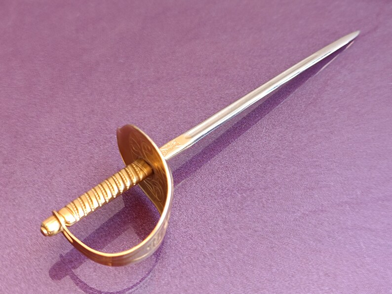K&ouml;nnte beinhalten: Ein goldfarbener Metall-Cocktail-Stich in Schwertform mit einem gedrehten Griff und einer dekorativen Schutzvorrichtung. Der Stift liegt auf einer violetten Oberfl&auml;che.