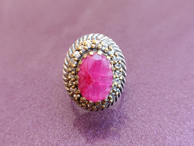 Op de afbeelding: Een zilveren ring met een roze edelsteen omringd door kleinere witte edelstenen. De ring heeft een getextureerde band.