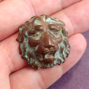 Pode incluir: Um botão metálico circular com uma escultura em relevo de uma cabeça de leão. A superfície apresenta um tom castanho-cobre quente com oxidação verde visível nas áreas rebaixadas da juba e nos traços faciais, realçando a textura.