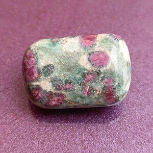 Puede incluir: Una piedra rodada de forma rectangular con bordes redondeados. La piedra tiene una base verde con manchas rojas y blancas. Las manchas rojas se concentran en la superficie. Los colores son una mezcla de verde, rojo y blanco.