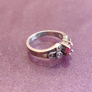 K&ouml;nnte beinhalten: Ein silberner Ring mit einem runden, leuchtend roten Edelstein. Der Ring hat dekorative Details mit kleinen, dunklen Akzenten um den Edelstein und am Band. Der Ring ist vor einem lila Hintergrund platziert.