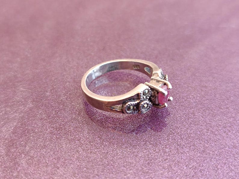 K&ouml;nnte beinhalten: Ein silberner Ring mit einem runden, leuchtend roten Edelstein. Der Ring hat dekorative Details mit kleinen, dunklen Akzenten um den Edelstein und am Band. Der Ring ist vor einem lila Hintergrund platziert.