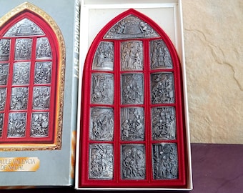May include: A silver-colored religious relief set in a red arched frame, displayed in a white box. The relief is divided into rectangular panels depicting biblical scenes. The box is labeled "RETAULE VALENCIA DE NADAL".