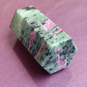 May include: A polished, hexagonal ruby zoisite crystal. The stone features a mottled pattern of green, black, and pink hues. The crystal is displayed on a purple surface, showcasing its natural beauty.