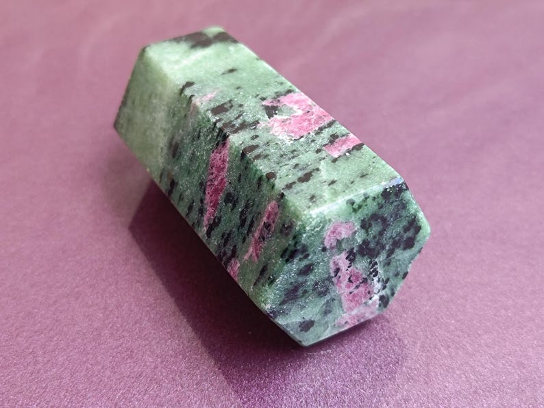 May include: A polished, hexagonal ruby zoisite crystal. The stone features a mottled pattern of green, black, and pink hues. The crystal is displayed on a purple surface, showcasing its natural beauty.