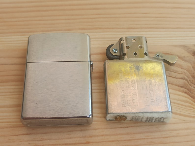 K&ouml;nnte beinhalten: Zwei silberne Feuerzeuge, eines mit geb&uuml;rsteter Oberfl&auml;che und das andere mit goldener Oberfl&auml;che. Das goldene Feuerzeug hat einen kleinen Hebel an der Seite und Text auf der Vorderseite.