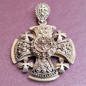 May include: A silver pendant in the shape of a cross with intricate details and a floral design. The cross is made up of four smaller crosses.
