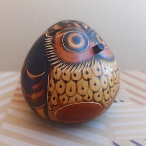 Puede incluir: Una figurita de búho de madera tallada a mano, pintada en marrón y negro, con un diseño detallado de plumas y ojos. El búho tiene un pequeño pico de madera y está firmado "D.SNARK".