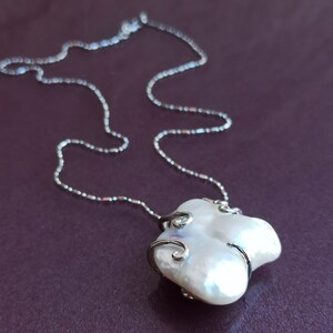 May include: A silver chain necklace with a white baroque pearl pendant. The pearl is set in a silver frame with delicate swirls.
