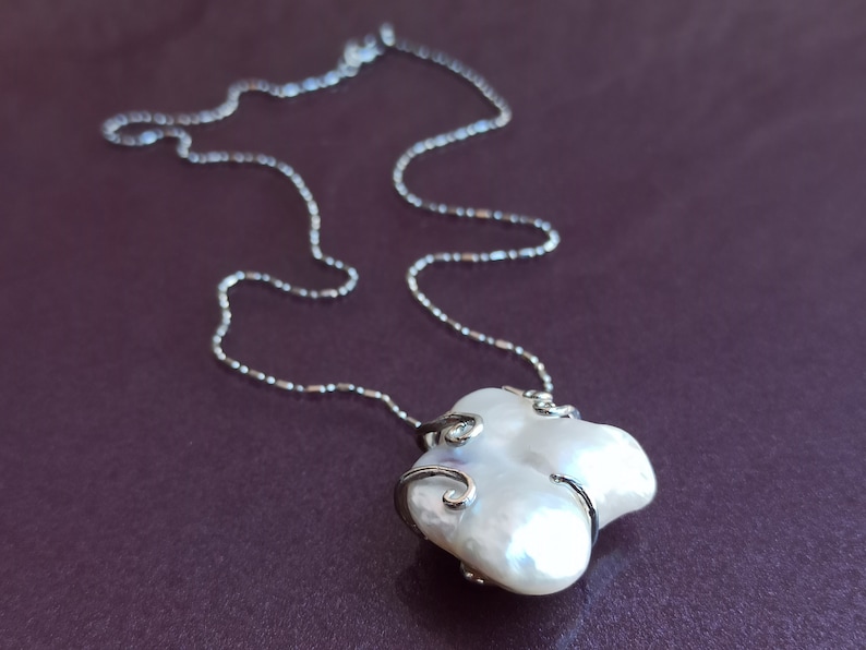 May include: A silver chain necklace with a white baroque pearl pendant. The pearl is set in a silver frame with delicate swirls.