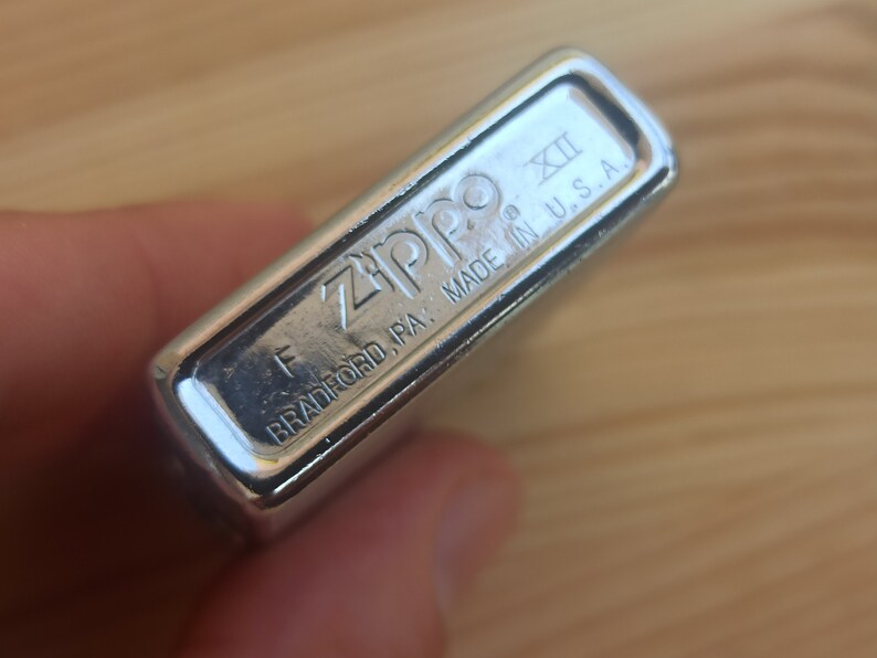 K&ouml;nnte beinhalten: Nahaufnahme eines silbernen Zippo-Feuerzeugs mit dem Text "Zippo" und "Bradford, PA. Made in U.S.A." auf der Unterseite. Das Feuerzeug hat eine kleine "XII" auf der Seite eingraviert.