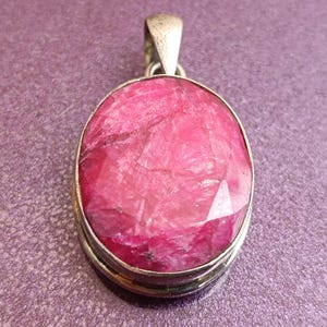 May include: A silver-coloured pendant featuring a large, oval-cut, pink gemstone. The gemstone is set within a metal frame and has a loop for a chain. The background is a textured, purple surface.