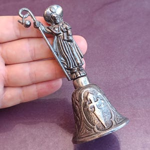 May include: A silver-toned metal hand bell featuring a sculpted figure of a robed person holding a staff with a curved top. The bell base is decorated with an embossed cross design and intricate scrollwork patterns on the exterior surface.