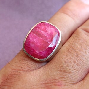 Op de afbeelding: Een ring met een grote, rechthoekige robijn edelsteen in een zilveren zetting. De robijn heeft een levendige roze kleur en een gefacetteerd oppervlak. De ring wordt aan een vinger getoond, waarbij het ontwerp en de kleur van het sieraad worden getoond.