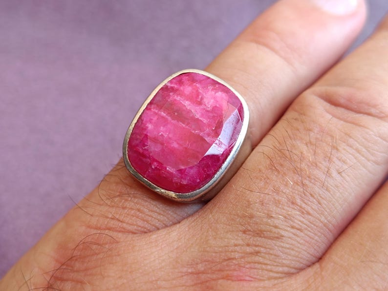 Op de afbeelding: Een ring met een grote, rechthoekige robijn edelsteen in een zilveren zetting. De robijn heeft een levendige roze kleur en een gefacetteerd oppervlak. De ring wordt aan een vinger getoond, waarbij het ontwerp en de kleur van het sieraad worden getoond.