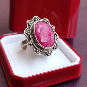 Op de afbeelding: Een zilveren ring met een grote, facetgeslepen ovale robijn edelsteen. De ring heeft een gedetailleerde, sierlijke zetting met een geschulpte rand en kralenaccenten. Gepresenteerd in een rode sieradendoos met een wit interieur.