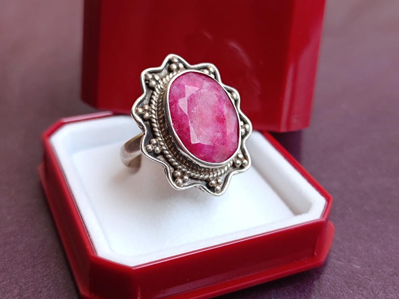 Op de afbeelding: Een zilveren ring met een grote, facetgeslepen ovale robijn edelsteen. De ring heeft een gedetailleerde, sierlijke zetting met een geschulpte rand en kralenaccenten. Gepresenteerd in een rode sieradendoos met een wit interieur.