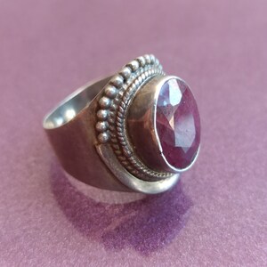 Pode incluir: Um anel de prata com uma grande gema de rubi facetada de forma oval, engastada em um engaste de bezel. A banda do anel &eacute; decorada com um design de contas.