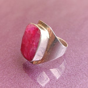 May include: A silver ring with a large, rectangular, faceted ruby gemstone. The ruby is a vibrant pinkish-red. The ring has a modern design with a wide band and a bezel setting. A statement piece of jewellery.