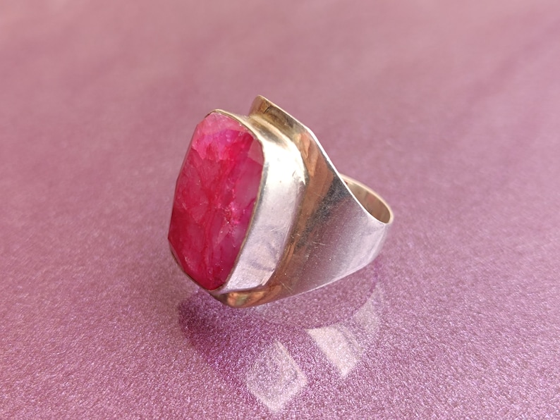 May include: A silver ring with a large, rectangular, faceted ruby gemstone. The ruby is a vibrant pinkish-red. The ring has a modern design with a wide band and a bezel setting. A statement piece of jewellery.