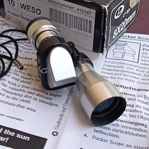 Peut inclure: Un WESO Pocket Scope, un petit monoculaire noir et argenté, est posé sur un document imprimé. L'appareil est doté d'un cordon noir et est accompagné de sa boîte d'origine. Le texte "Pocket Scope" est visible.