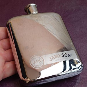 Pode incluir: Uma petaca retangular de metal prateado com topo arredondado e tampa de rosca. A superfície frontal apresenta o texto gravado Jameson Irish Whiskey e um logótipo circular com as letras JJ&S e John Jameson & Son Limited.
