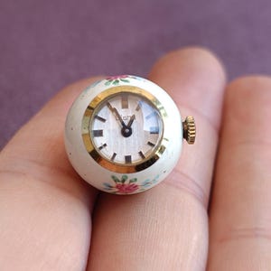 May include: A small, round vintage watch with a white face and gold-tone bezel. The watch has a white body with floral accents in pink, blue, and green. The watch is approximately 1 inch in diameter.