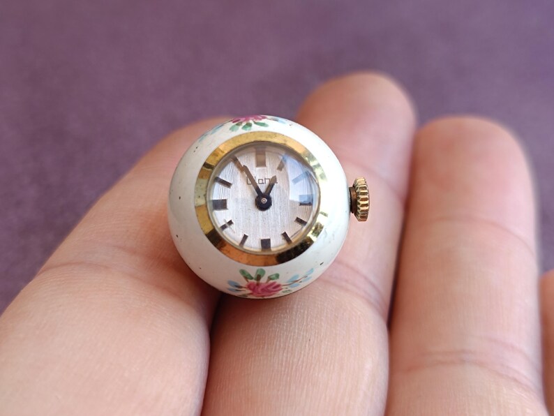 May include: A small, round vintage watch with a white face and gold-tone bezel. The watch has a white body with floral accents in pink, blue, and green. The watch is approximately 1 inch in diameter.