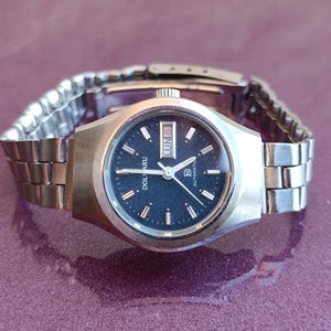 May include: A vintage silver-toned wristwatch with a dark blue face. The watch features a day and date display, with the day reading "LUN 6". The watch has a silver metal band and is set against a purple background.