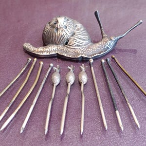 Peut inclure: Une figurine d'escargot en métal argenté avec une coquille texturée et des antennes étendues, accompagnée de dix épingles métalliques fines. Trois épingles présentent de petits embouts texturés en forme de bulbe, les sept autres sont lisses.
