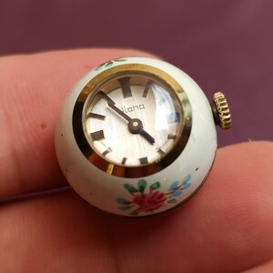 May include: A small, round vintage watch with a white face and gold-tone accents. The watch face has the word "lana" printed on it. The watch is decorated with pink and blue floral designs. The watch's crown is gold-toned.
