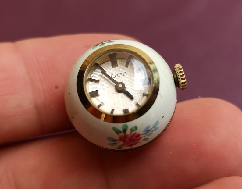 May include: A small, round vintage watch with a white face and gold-tone accents. The watch face has the word "lana" printed on it. The watch is decorated with pink and blue floral designs. The watch's crown is gold-toned.