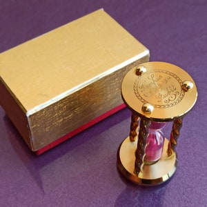 May include: A gold hourglass with a pink sand timer. The base of the hourglass is engraved with the text "Zumphrus & Co.  Frankfurt".