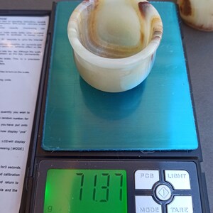 K&ouml;nnte beinhalten: Ein wei&szlig;es und braunes Onyx-Schnapsglas mit einem Gewicht von 7,137 Gramm auf einer digitalen Waage.