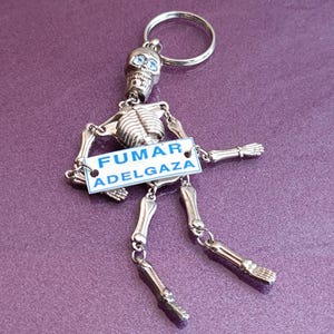 May include: A silver skeleton keychain with a blue and white sign that reads "FUMAR ADELGAZA".