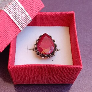 Pode incluir: Um anel de pedra vermelha em uma caixa de anel de veludo vermelho. O anel tem uma pedra em forma de l&aacute;grima com uma configura&ccedil;&atilde;o de prata.