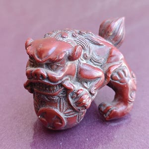 May include: A small, red, wooden sculpture of a mythical lion-like creature with intricate details. The creature is holding a round object in its paws.