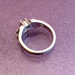 K&ouml;nnte beinhalten: Ein silberner Ring mit einem zentralen rosa Edelstein und kleineren Akzentsteinen. Der Ring ist vor einem lila Hintergrund platziert, der sein kompliziertes Design und seine Details hervorhebt. Das Band des Rings ist glatt und poliert.
