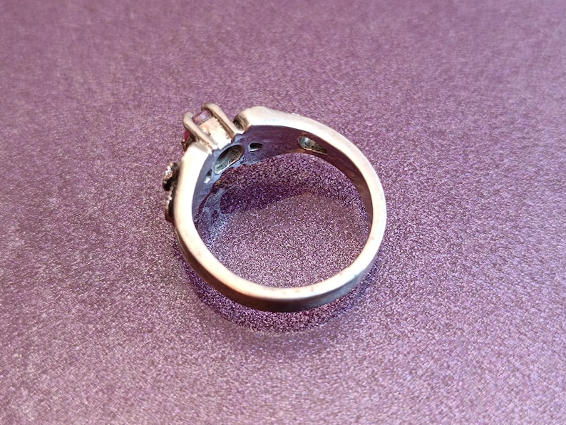 K&ouml;nnte beinhalten: Ein silberner Ring mit einem zentralen rosa Edelstein und kleineren Akzentsteinen. Der Ring ist vor einem lila Hintergrund platziert, der sein kompliziertes Design und seine Details hervorhebt. Das Band des Rings ist glatt und poliert.