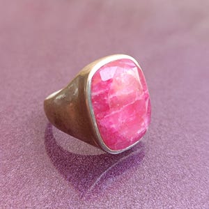 Op de afbeelding: Een zilveren ring met een grote, facetgeslepen, rechthoekige roze edelsteen. De ring heeft een dikke band en een bezel setting die de steen vasthoudt. De edelsteen heeft een gemarmerd uiterlijk met variaties in de roze kleur.