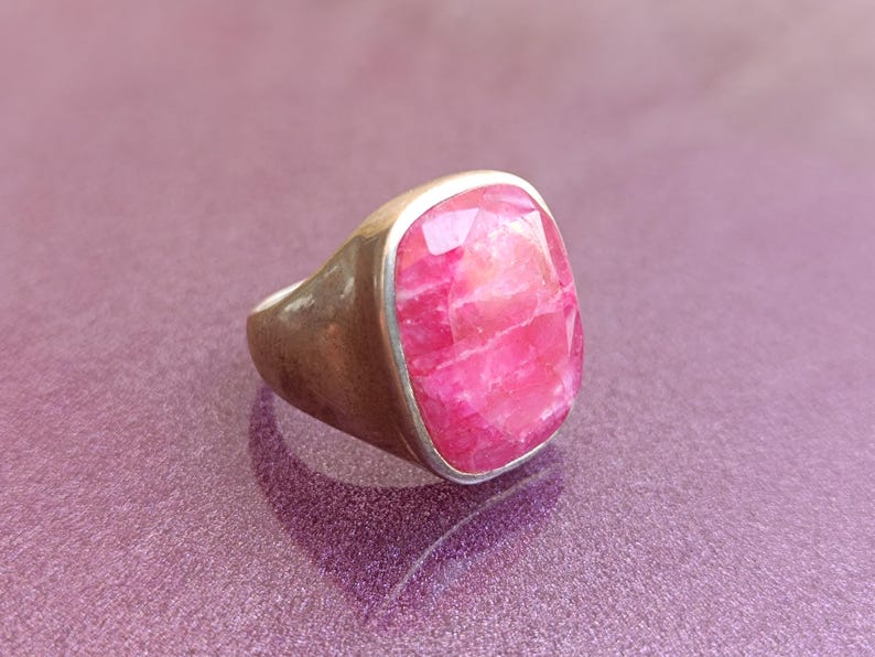 Op de afbeelding: Een zilveren ring met een grote, facetgeslepen, rechthoekige roze edelsteen. De ring heeft een dikke band en een bezel setting die de steen vasthoudt. De edelsteen heeft een gemarmerd uiterlijk met variaties in de roze kleur.