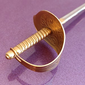 K&ouml;nnte beinhalten: Ein dekoratives Schwert mit goldenem Griff und silberner Klinge. Der Griff ist mit einem detaillierten Muster verziert. Das Schwert liegt auf einer violetten Oberfl&auml;che.