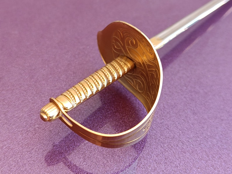 K&ouml;nnte beinhalten: Ein dekoratives Schwert mit goldenem Griff und silberner Klinge. Der Griff ist mit einem detaillierten Muster verziert. Das Schwert liegt auf einer violetten Oberfl&auml;che.