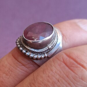 Pode incluir: Um anel de prata com uma grande gema oval rosa engastada em um engaste de bezel. O anel tem uma banda decorativa com uma borda de contas.